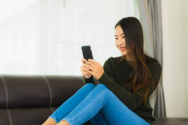 Güzel bir Asyalı kadın oturma odasındaki koltukta akıllı cep telefonu ya da cep telefonu kullanıyor.