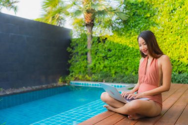 Genç Asyalı bir kadının portresi oteldeki yüzme havuzunda dizüstü bilgisayar kullanıyor.