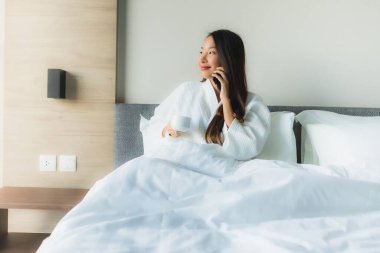 Güzel Asyalı genç kadınların portresi. Mutlu bir gülümseme. Yatak odasındaki yatakta kahve ve cep telefonuyla.