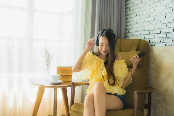 年轻的亚洲女子坐在椅子上 一边听音乐 一边用手机喝咖啡 一边在客厅里看书 — 图库照片