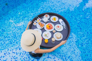 Asyalı güzel bir kadının portresi yüzme havuzunda yüzen kahvaltı tepsisiyle mutlu bir gülümseme