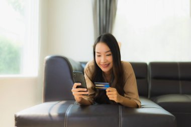 Akıllı telefon ya da kredi kartıyla oturma odasında online alışveriş yapan Asyalı güzel bir kadının portresi.