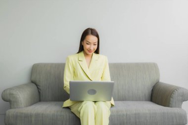 Portre güzel genç Asyalı kadın oturma odasındaki koltukta dizüstü bilgisayar kullanıyor.