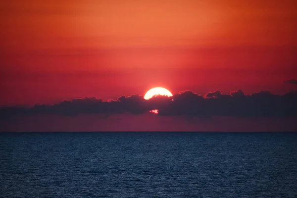 Dramatic sunset over the ocean horizon at dusk with clouds partly covering the sun against a deep red sky background - dreamy glowing light fading into the waters