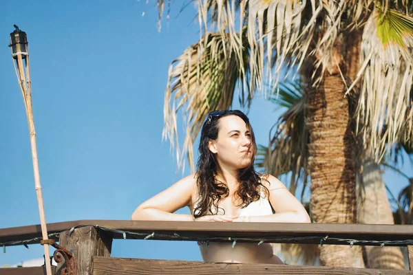 An attractive young woman in a white bikini leaning on a balcony railing in front of a palm tree looking into the distance