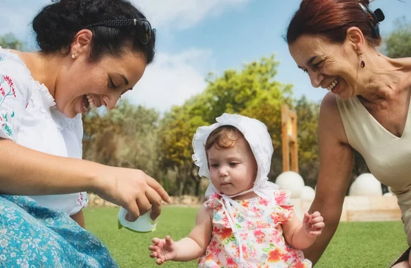 A happy family of 3 generations, baby, mother and grandmother sitting on the grass and playing outdoors in a public park with a blue sky background