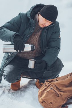 Kış ormanlarında, ceketli ve turuncu çizmeli bir adam termostan kahve içiyor. Yüksek kalite fotoğraf