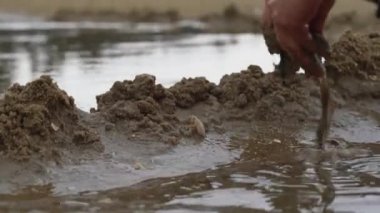 Bir insan kumsalda deniz kenarında kum yığınları inşa ediyor. Kum heykeli barajı inşa ediyor. Kum heykeli barajından akan suya yakın çekim yapıyor.