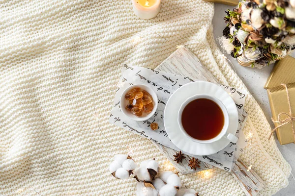 Cozy winter composition with hot tea in a cup. a Cup of black tea on wooden tray on knitted plaid with candles, presents and Christmas tree. Christmas menu, winter tea party. Flat lay, copy space