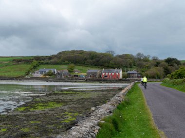 West Cork, İrlanda, 2 Mayıs 2022. Bulutlu bir günde kıyıda küçük bir yerleşim yeri. Alglerle kaplı deniz yatağı alçak gelgitte. Clonakilty Körfezi yakınlarında bir İrlanda köyü. Bisikletçiler.
