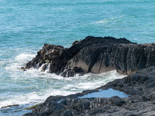 Tidal waves break on black coastal rocks, seascape. The water is turquoise in color. White foam on the waves, splashes, rock beside sea.
