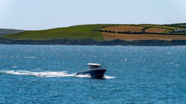 County Cork, İrlanda, 28 Mayıs 2022. Güneşli bir bahar gününde mavi bir denizde beyaz ve mavi bir motorlu tekne yol alıyor. Deniz Burnu.