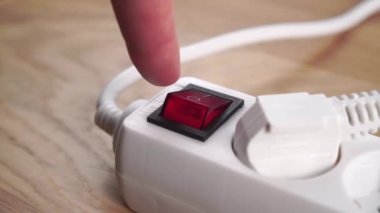Parmak, beyaz bir elektrik uzatma kablosunu kapatıyor. Düğmeye yakın basıyor. Elektrik tasarrufu kavramı. Kırmızı düğme. Bağlı elektrik prizi. Ahşap bir arka planda