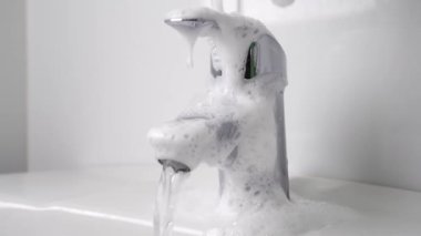 Banyo temizliği bitti. Koruyucu eldivendeki bir el, musluğu deterjan köpüğüyle yıkar, süngeri yavaş çekimde sıkar. Akan su. Hijyenik ovma. Ödev konsepti