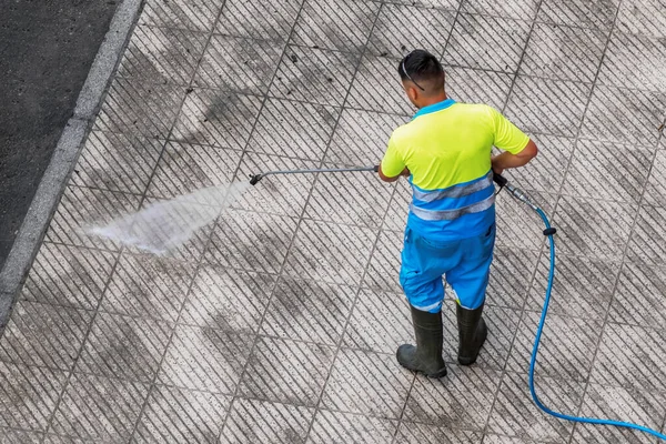 Street washer man washing dirty street pavement with high pressure water machine. Worker in uniform and rubber boots cleaning city sidewalk