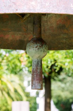 Heavy antique bell clapper against the background of summer foliage of trees. Textured rusty iron surface. Historical landmark