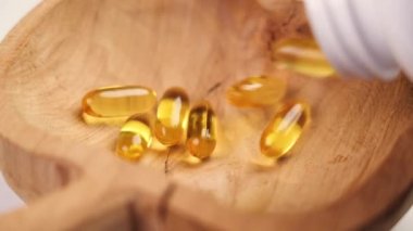 SağIık desteği. Sarı Omega 3 kapsülleri beyaz bir plastik şişeden kırık bir tahta kaseye düşüyor. Kırsal bakış açısı. Yavaş çekim