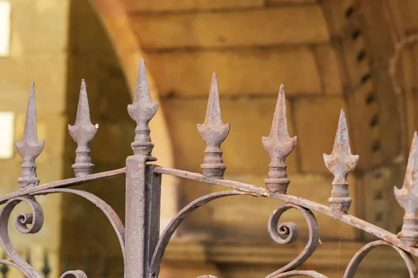 Rusty forged iron fence with spikes and peaks of an antique medieval castle with a brown stone wall in blur. Ancient spear shape