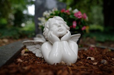 Mezarda yatan küçük beyaz melek figürü bulanık arka plan çiçekleri ve mezar taşına bakıyor.