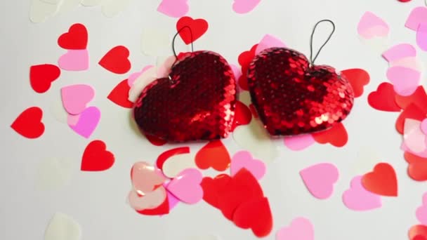 Два красных сердца из блёсток и конфетти от разноцветных сердец раскачиваются на белом фоне — стоковое видео