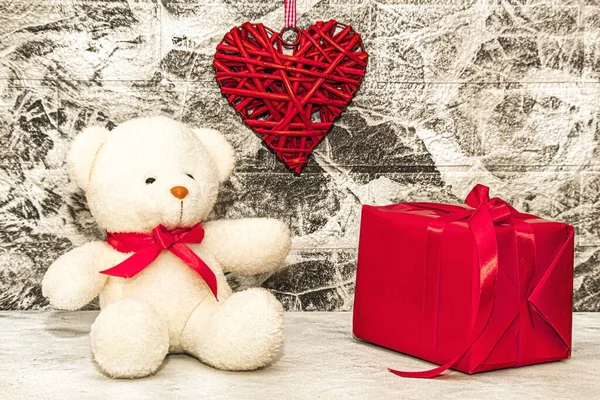 White Teddy bear with red heart and gift box on grey wall background. Rattan work of heart shape