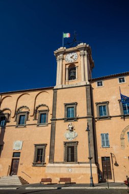 6 Şubat 2022 - Tarquinia, Viterbo, Lazio, İtalya - Köyün ana meydanı. Belediye binası güneşle aydınlandı, mavi gökyüzünde. Saati olan kule. İtalyan bayrağı.