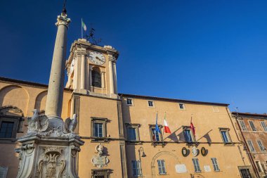 6 Şubat 2022 - Tarquinia, Viterbo, Lazio, İtalya - Köyün ana meydanı. Dikilitaş ve haçlı çeşme. Belediye binası ve saatli kule. İtalyan bayrağı. Güneş ve mavi gökyüzü.