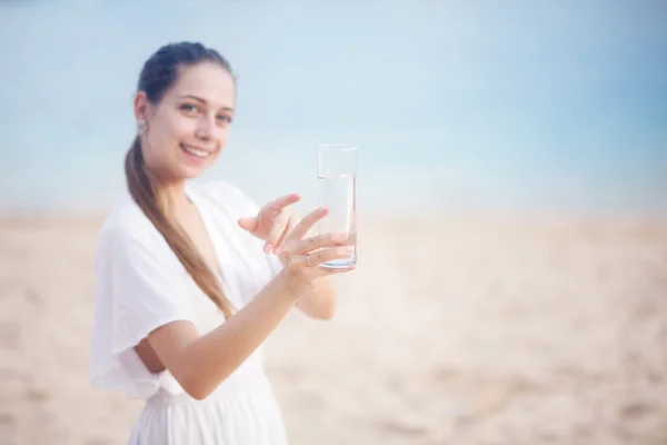 Mujer joven en la orilla del océano en vestido blanco con vaso de agua — Foto de Stock