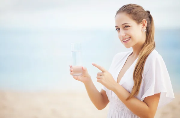 Jovem mulher na costa do oceano em vestido branco com vidro de água — Fotografia de Stock