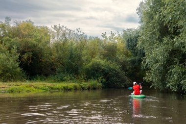A narrow river among thickets of trees and bushes.. Girl surfer in red t-shirt sitting on the sup board. Paddleboard SUP Water Sport Activity Holidays Vacation. Copy space.