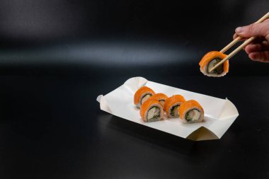 A cardboard box for sushi, a man hand using chopsticks takes a roll. Black background. Place for text. Asian cuisine concept. White paper box for food products. Cardboard crafts. Copy space.