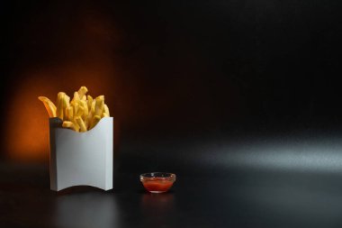 Kartondan yapılmış beyaz kağıt ambalajı. Kızarmış patates kızartması, konteynırda. Fast food, üretim konsepti. Siyah arka plan. Soslu küçük cam bir tabak. Uzayı kopyala Sarı arka ışık