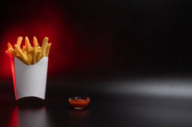 Kartondan yapılmış beyaz kağıt ambalajı. Kızarmış patates kızartması, konteynırda. Fast food, üretim konsepti. Siyah arka plan. Soslu küçük cam bir tabak. Boşluğu kopyala.