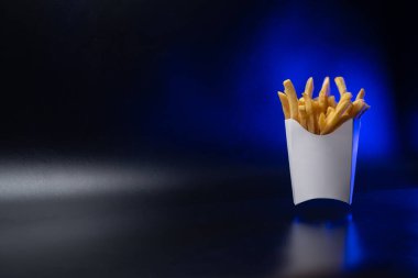 Kartondan yapılmış beyaz kağıt ambalajı. Kızarmış patates kızartması, konteynırda. Fast food, üretim konsepti. Siyah arka plan. Metin eklemek için bir yer. Uzayı kopyala Mavi arka ışık