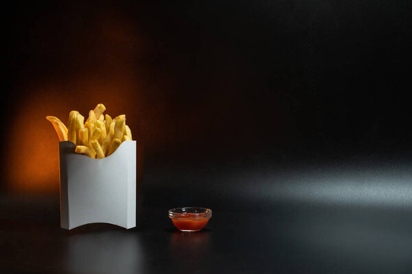 Упаковка из белого картона. Картошка фри, жареная, в контейнере. Фастфуд, концепция производства. Чёрный фон. Маленькая стеклянная тарелка с соусом. Принято. Желтая подсветка