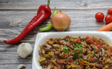 Biber ve domates, soğan, sarımsak ve zeytinyağıyla sotelenmiş mantar, beyaz bir tabakta, ahşap arka planda..