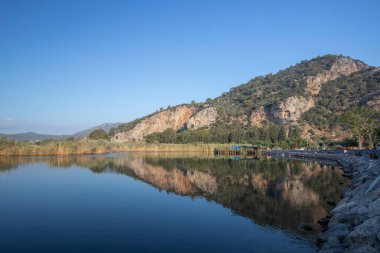 The rock-cut temple tombs of the ancient city of Kaunos in Dalyan, Mula, Turkey. Beautiful view of Dalyan river with reed beds, excursion boats and carved tombs in the background.