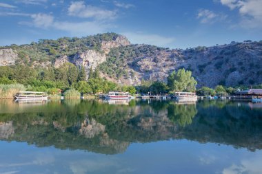 The rock-cut temple tombs of the ancient city of Kaunos in Dalyan, Mula, Turkey. Beautiful view of Dalyan river with reed beds, excursion boats and carved tombs in the background.