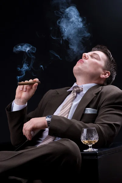 Young businessman sitting on a couch with an alcoholic drink and a cigar — Stock Photo, Image