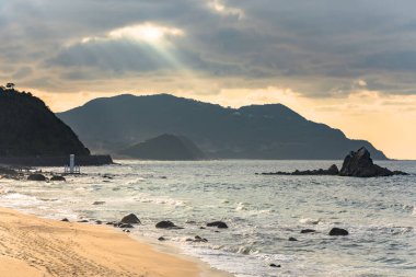 The sunlight filtering through the clouds at dusk on the Sunset Beach leading to the Sakurai Futamigaura's Meoto Iwa Couple Rock protected by a sacred white Shinto torii gate in the sea off Fukuoka.