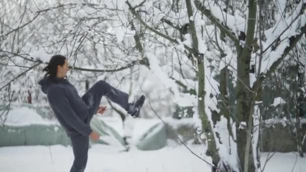 Atleta masculino, con el pelo negro largo, patea un árbol, la nieve está cayendo sobre él — Vídeos de Stock