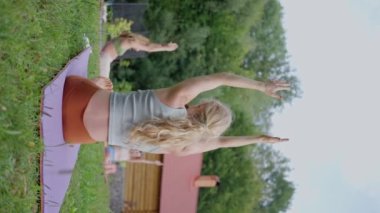 Ön planda arkadaşlarıyla birlikte sarışın bir kadın, yoga yapıyor, yazın çimlerin üzerinde egzersiz yapıyor, yavaş çekim, güneşli bir gün, dikey görüş. Yüksek kalite 4k görüntü