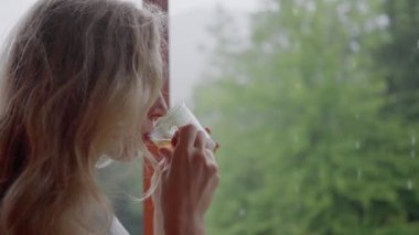 Uzun beyaz saçlı kadın, balkonda duruyor ve sıcak çay içiyor, dışarıda yağmur yağıyor, dikey video. Yüksek kalite 4k görüntü