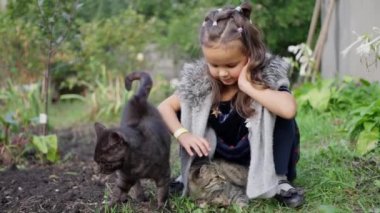 Küçük kız bahçede yatan gri bir kediyi öpüyor, kara kedi etrafta dolaşıyor, yaramaz ve neşeli kedi, kız kedilerle oynuyor. Ağır çekim. Yüksek kalite 4k görüntü