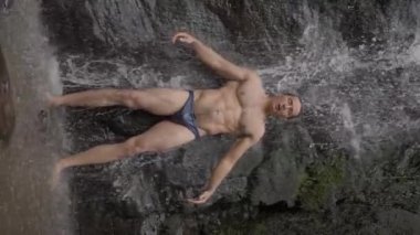 Çıplak gövdeli, uzun siyah saçlı, seksi bir erkek sporcu, bir dağ nehrinde şelalenin altında duruyor. Ağır çekim. Dikey video. Yüksek kalite 4k görüntü