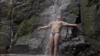 Çıplak gövdeli, uzun siyah saçlı, seksi bir erkek sporcu, bir dağ nehrinde şelalenin altında duruyor. Yavaş hareket. Yüksek kalite 4K görüntü.