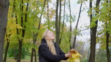 Neşeli güzel kadın sonbahar parkına sarı yapraklar fırlatıyor. Ağır çekim. Genç kızlar sonbahar akçaağaç yaprakları kusar. Neşeli duygular, genç kız için güzel bir gün..