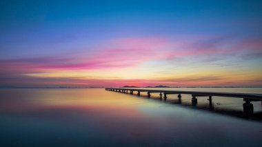 Mar Menor, Cartagena, İspanya 'da gün doğumunda güzel bir manzara. Göz kamaştırıcı, renkli bir gökyüzü ve denizin sakin sularına yansıyan. Ayrıca suda ahşap bir iskele görüyoruz. Huzur ve sükuneti iletir..
