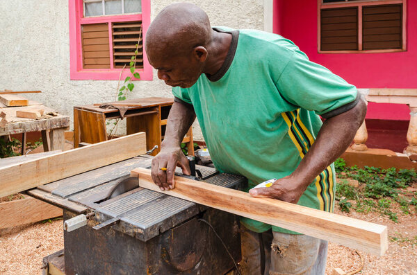 A cabinet maker with a pencel between his fingers bends over the table of an old electric saw as he supports a piece of 2 x 2 wood that he uses the saw to cut its end.
