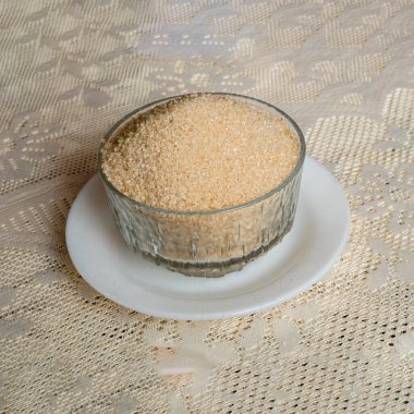 Altın kahverengi şekerle dolu şeffaf çanak çömlekli beyaz bir çay tabağı fildişi renkli masa örtüsüyle kaplı bir masada..